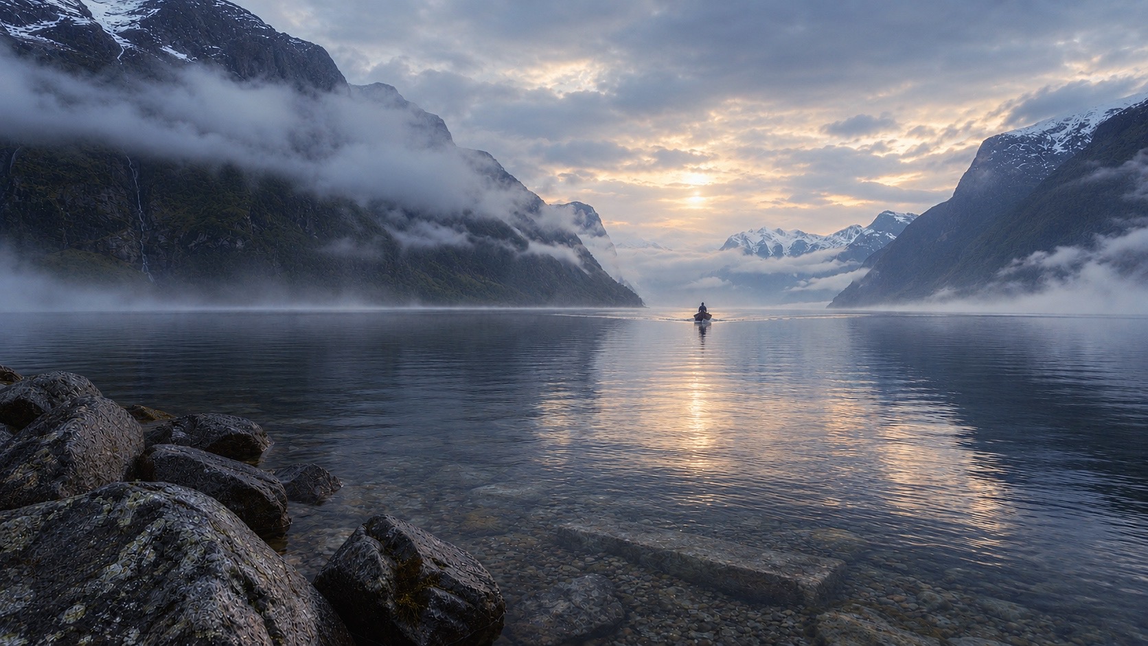 Ein Mensch in einem kleinen Boot auf einem stillen Fjord, im Morgenlicht zwischen Nebel und Bergen